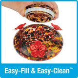 hand lifting the glass lid off of the base dish for easy fill and easy clean on the Nature's Way red, yellow and black speckled hand blown glass garden hummingbird feeder
